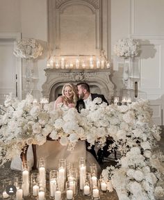 a bride and groom sitting in front of a fireplace surrounded by white flowers with candles