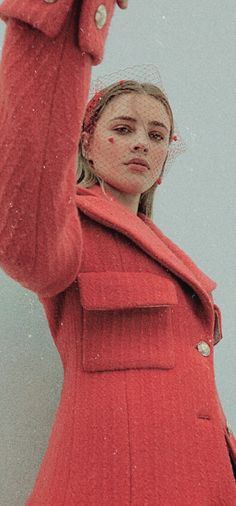 a woman in a red coat is holding her hand up to the side and looking off into the distance