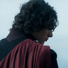 a man with curly hair wearing a cape and looking off into the distance on a cloudy day