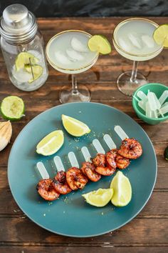 a blue plate topped with shrimp skewers next to two margarita glasses and lime wedges