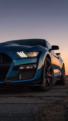 the front end of a blue car parked in a parking lot at sunset with its lights on