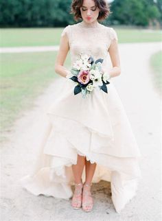 a woman in a white dress holding a bouquet and standing on a dirt road with trees in the background