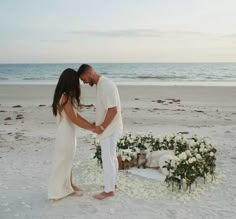 a man and woman standing on top of a sandy beach next to an arch covered in flowers