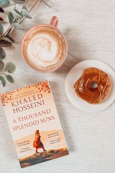 a cup of coffee next to a book and doughnut on a white tablecloth