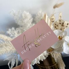 a person holding up a pink business card in front of a vase with white flowers