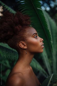 a woman with an afro haircut standing in front of some green plants and looking off into the distance