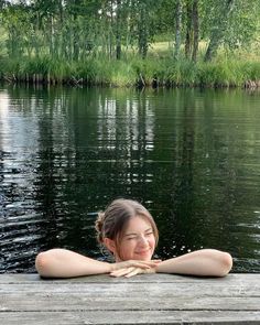 a woman sitting on top of a wooden dock next to a body of water with trees in the background