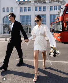 a man and woman walking across a parking lot holding each other's hands as they walk towards a red double decker bus