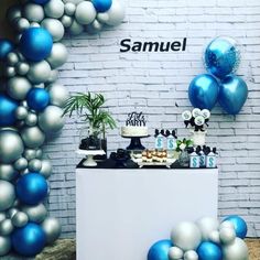 a table topped with blue and silver balloons next to a white brick wall filled with cake