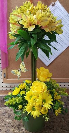 a vase filled with yellow flowers sitting on top of a stone floor next to a bulletin board