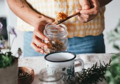 a person holding a spoon over a jar filled with dirt and flowers on top of a table