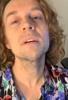 a man with long curly hair wearing a floral shirt and looking off into the distance