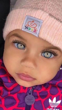 a baby with blue eyes wearing a pink hat and purple jacket is looking at the camera