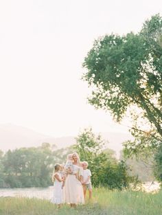 three children standing in the grass near a tree and water with mountains in the background