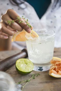 a person holding a lemon wedge over a glass filled with water and limes on a wooden table