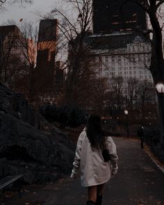 a woman walking down a path in a park at dusk with tall buildings behind her