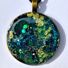 a round pendant with green and blue designs on it's side, sitting on a white surface