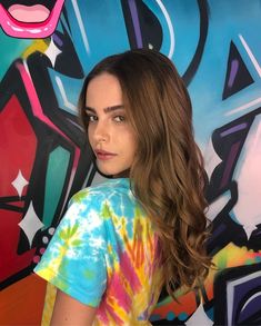 a woman with long hair standing in front of a colorful wall and wearing a tie dye shirt