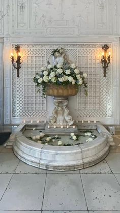 a fountain with flowers and candles on it in the middle of a room filled with white walls