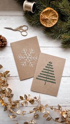 two christmas cards sitting on top of a table next to some gold foil and pine cones