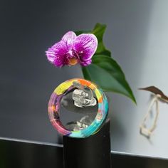 a glass vase with a flower in it on a table next to a purple flower