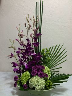 a bowl filled with purple and green flowers on top of a white tablecloth next to a wall