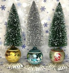 three small christmas trees in vases with snowflakes on the wall behind them