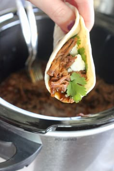 a hand holding a tortilla over a slow cooker filled with meat and vegetables