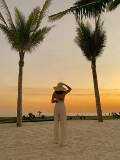 a woman standing on top of a sandy beach next to palm trees at sun set