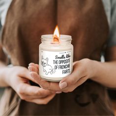 a person holding a jar with a candle in it that says, smell me the opposite of frenchie farts