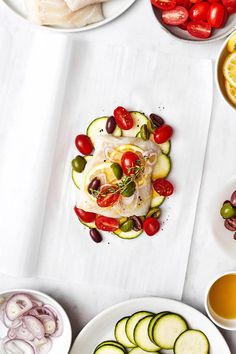 several plates of food are arranged on a white tablecloth with tomatoes, cucumbers, olives, and lemon slices