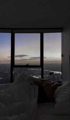 a person laying on a bed in front of a window with the city outside it