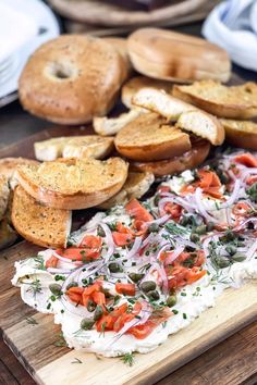 an assortment of breads and bagels on a wooden cutting board with tomatoes, onions, herbs