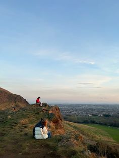 two people sitting on top of a hill next to a green field and blue sky