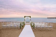 an outdoor wedding setup on the beach with white chairs and greenery draped over the aisle