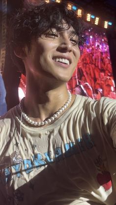 a young man with curly hair wearing a white t - shirt and pearl necklace smiling at the camera