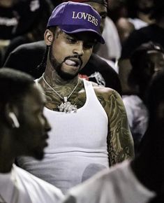 a man with tattoos wearing a purple hat and white tank top standing in front of other people