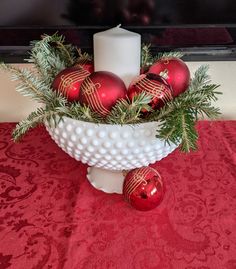 a white bowl filled with christmas ornaments and a candle on top of a red table cloth