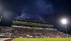 a stadium full of people watching a football game with lightning in the sky behind them