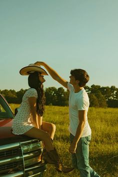 a man standing next to a woman sitting on the hood of a car in a field