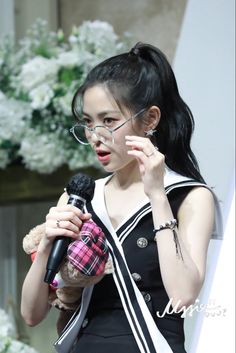 a woman holding a teddy bear while wearing glasses and speaking into a microphone in front of flowers