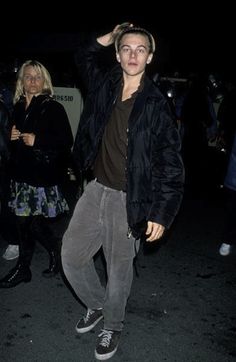 a man in black jacket and grey pants standing next to other people on street at night