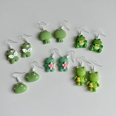 there are many green frog earrings on the white surface, one is wearing a pink bow