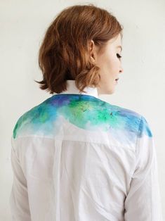 a woman wearing a white shirt with blue and green designs on the back, standing in front of a white wall