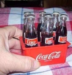 a hand is holding a small coca - cola box with six bottles in it on a checkered tablecloth