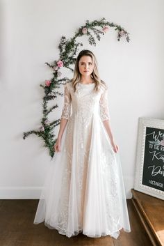 a woman wearing a white dress standing in front of a wall with flowers and greenery