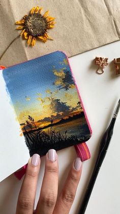 a woman's hand holding an open notebook with a painting on it