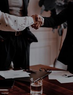 two people shaking hands over a desk with papers and glasses on it, while another person holds the other hand