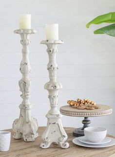 two white candles sitting on top of a wooden table next to a plate and cup