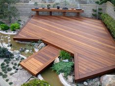 a wooden deck in the middle of a garden with plants and rocks around it, next to a pond